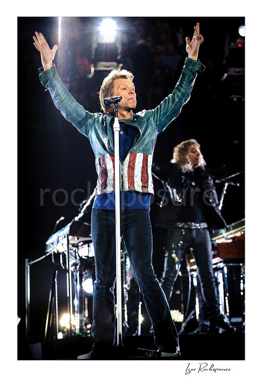 Vertical color image of Jon Bon Jovi on stage with both arms raised beside the microphone stand, with bright backlight and a blurred band member behind him