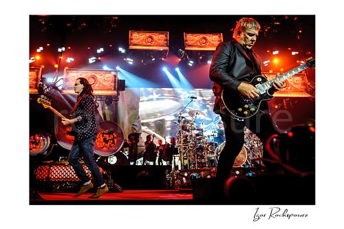 Horizontal color image of Rush performing live on stage with Geddy Lee, Neil Peart, and Alex Lifeson under vivid red and blue concert lighting