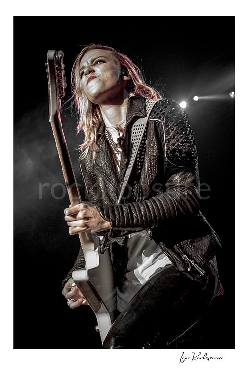 Vertical color image of Lzzy Hale of Halestorm playing guitar in a dramatic live performance close-up, with a studded leather jacket, stage smoke, and bright backlight behind her