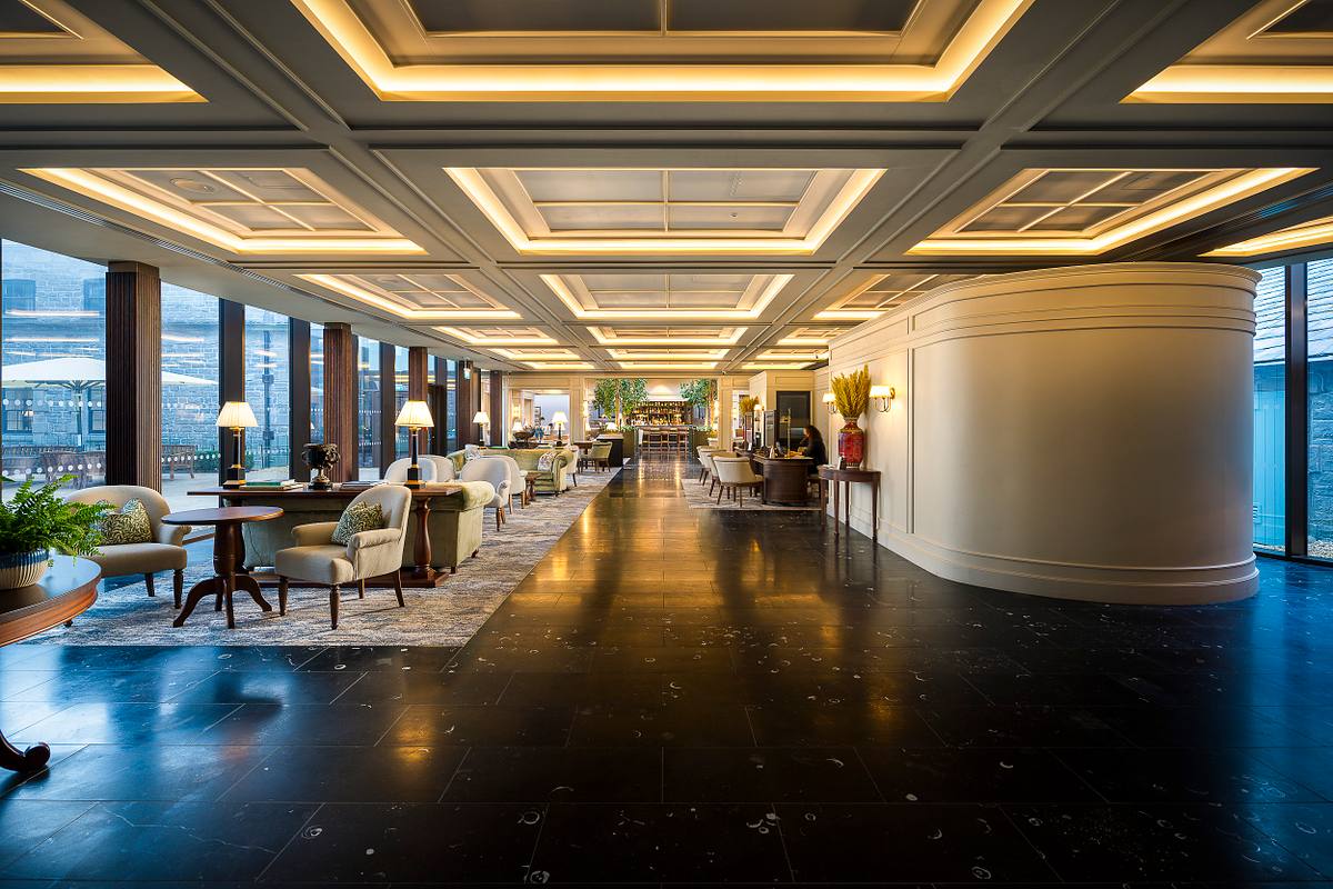 Interior view of the spacious, classically-styled lobby at Carton House, a Fairmont Managed Hotel, near Dublin. Features include a high, coffered ceiling with recessed lighting, dark polished floors, large windows, and multiple seating areas with armchairs and tables. A curved wall section is visible.