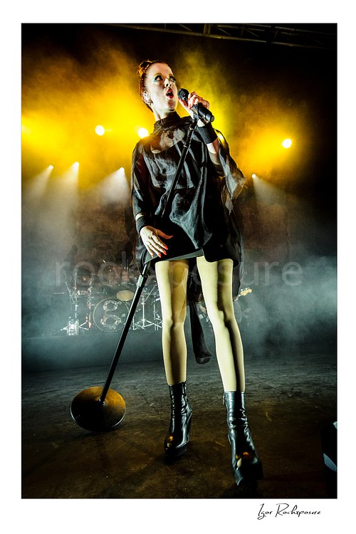 Vertical color image of Shirley Manson singing into a microphone during a Garbage concert, standing with a tilted mic stand in stage smoke under vivid yellow backlight