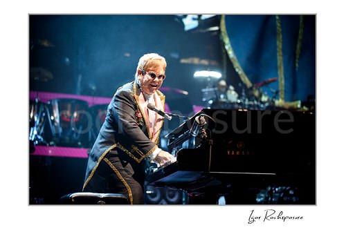 Horizontal color image of Elton John playing a Yamaha piano under blue stage lighting with a bright spotlight behind him
