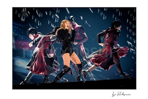 Horizontal color image of Taylor Swift singing on stage with dancers in sweeping coats under dramatic concert lighting