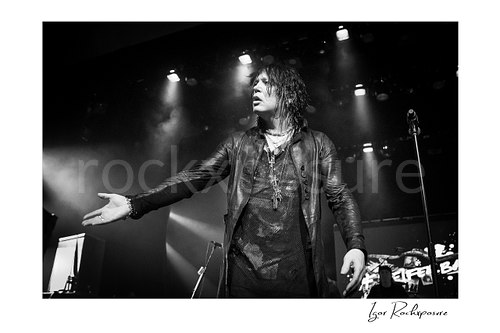 Horizontal black and white image of Tom Keifer on stage in a dramatic live performance moment, extending one hand outward beneath bright overhead spotlights with the microphone stand beside him