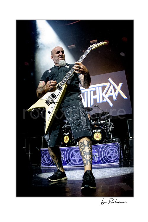 Vertical color image of Scott Ian of Anthrax performing live on stage, playing guitar in front of the Anthrax stage backdrop under bright concert lighting