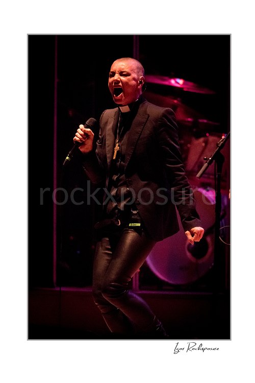 Sinéad O’Connor sings with knees bent and a microphone held near her chest, wearing a black jacket with a drum kit glowing pink behind her.