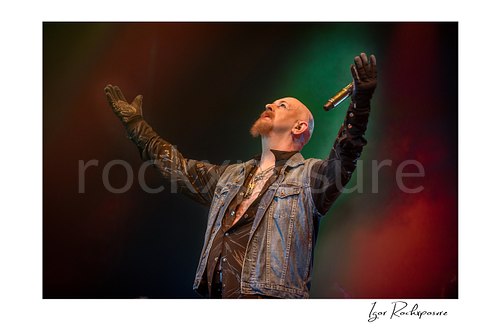 Horizontal color image of Rob Halford of Judas Priest performing live on stage with both arms raised, holding a microphone under dramatic red and green concert lighting