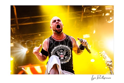 Horizontal color image of Ivan Moody of Five Finger Death Punch singing onstage with a microphone under intense yellow concert light