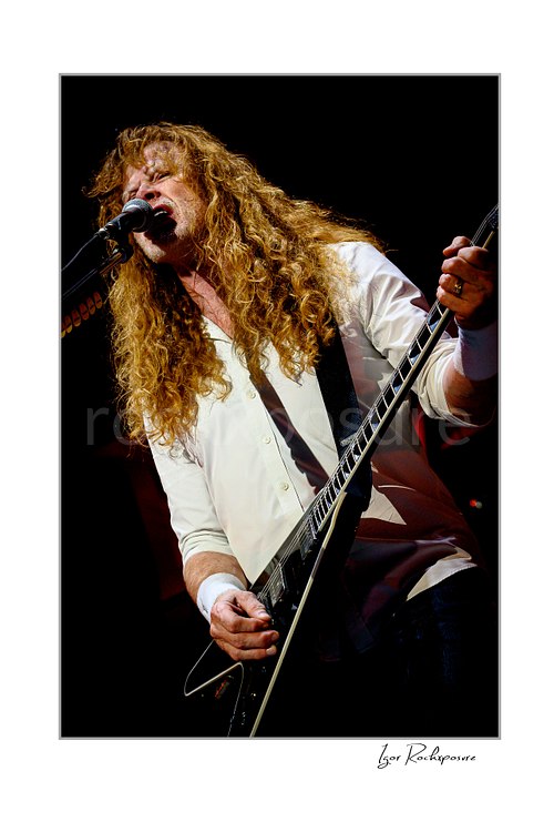 Vertical color image of Dave Mustaine of Megadeth singing into a microphone while playing guitar on stage with long curly hair