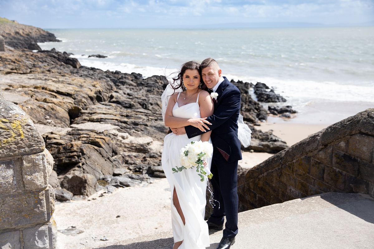 Bride and groom embracing above the rocky shoreline at Barry Island, South Wales