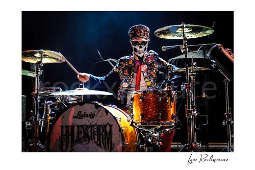 Horizontal color image of Arejay Hale of Halestorm playing drums in a dramatic live performance shot, wearing a skull face covering and a jacket covered in buttons behind a sparkling orange drum kit