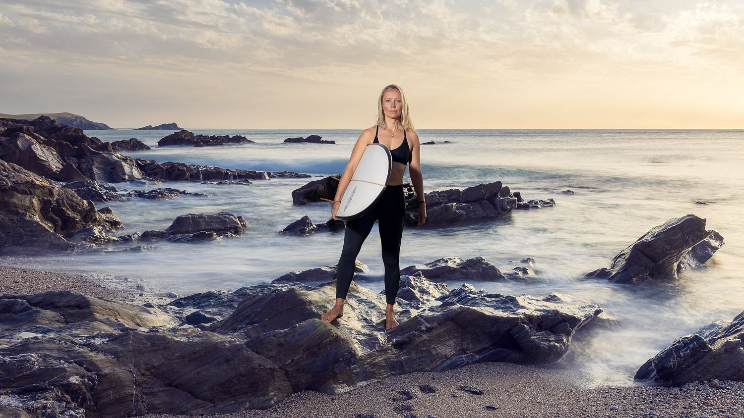 A woman in a wetsuit stands on a rocky shore at sunset, holding a surfboard, with waves and a cloudy sky in the background—capturing the perfect moment that any personal brand photographer would seek for an adventurous, coastal portfolio.