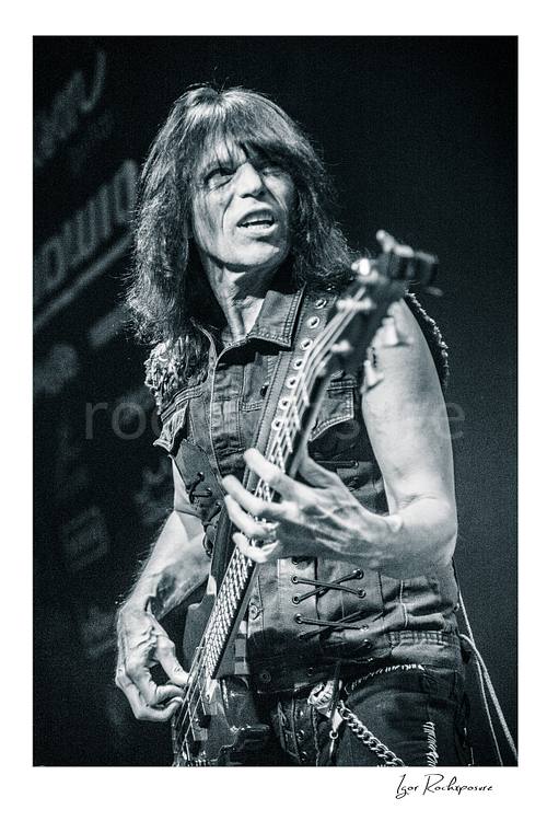 Vertical black and white image of Rudy Sarzo performing live with bass guitar in a close-up stage portrait