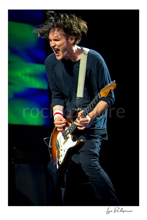 Josh Klinghoffer of Red Hot Chili Peppers plays a sunburst electric guitar, mouth open and hair flying above his face against green and blue stage color.