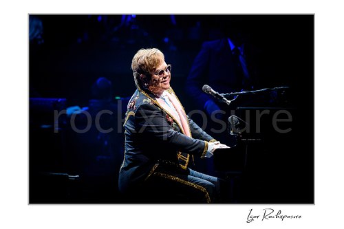 Horizontal color image of Elton John playing piano under deep blue stage lighting in a dark concert setting