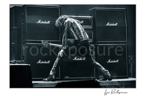 Horizontal black and white image of Nuno Bettencourt performing live on stage with an electric guitar before a wall of Marshall amplifiers