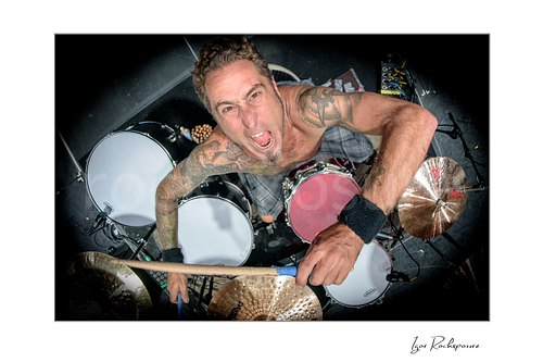 Horizontal color image of Sean Sellers of Beasto Blanco in an exclusive aerial overhead drummer shot, looking up at the camera with drumstick raised above the full drum kit