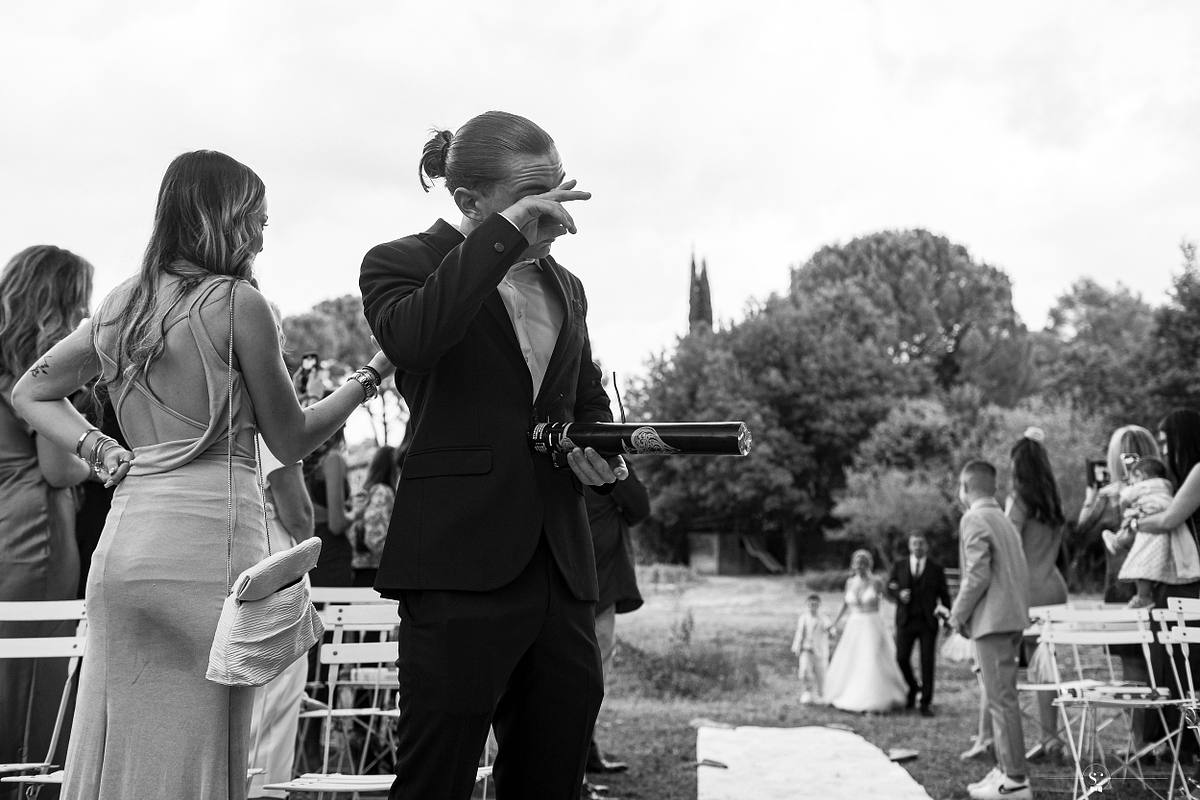 Un invit&eacute; masculin, les yeux couverts par la main, semble &eacute;mu, tandis qu'&agrave; l'arri&egrave;re-plan, des invit&eacute;s se retournent pour regarder la mari&eacute;e arriver, soulignant l'anticipation et l'&eacute;motion du mariage &agrave; N&icirc;mes
