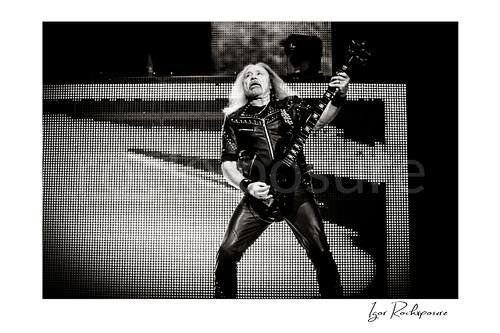 Horizontal black and white image of Glenn Tipton of Judas Priest performing live on stage, playing electric guitar in a wide stance against a bright LED screen backdrop