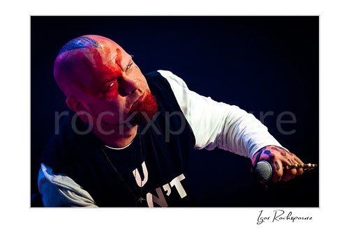 Horizontal color image of Ivan Moody of Five Finger Death Punch in a tight onstage close-up with microphone in hand under red and blue concert light