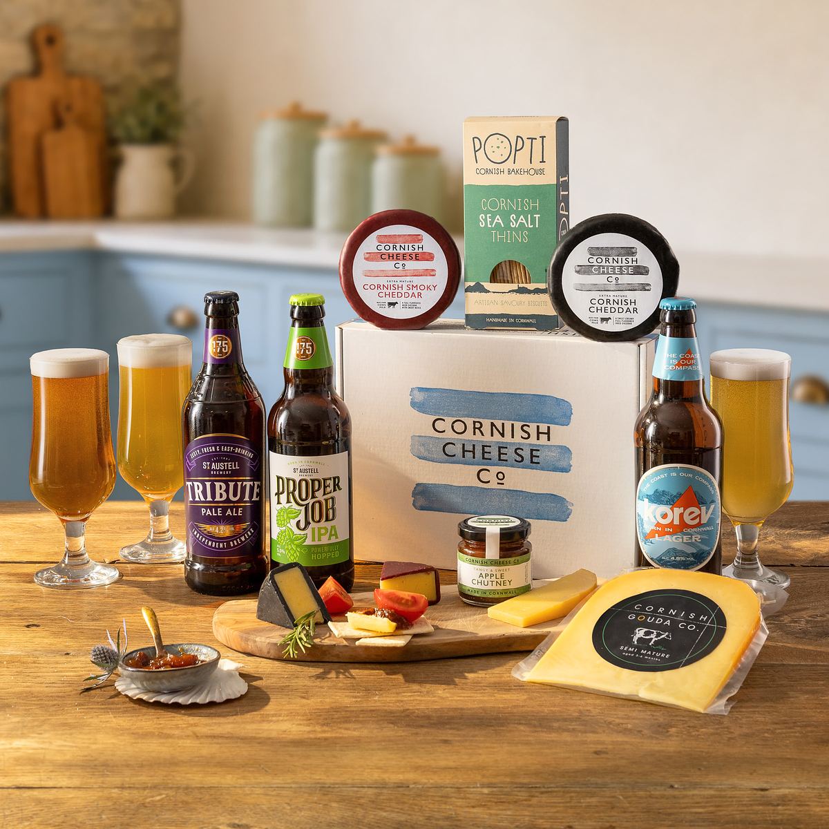 A selection of Cornish cheeses, crackers, and three bottled beers are artfully displayed on a table by a commercial photographer, with two filled beer glasses and kitchen items softly blurred in the background.