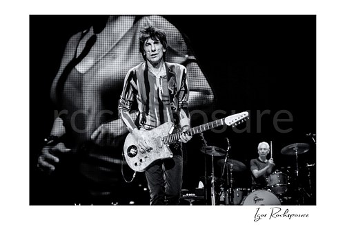 Horizontal black and white image of Ronnie Wood and Charlie Watts of The Rolling Stones performing on stage with electric guitar and drums