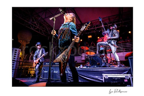 Horizontal color image of Jacob Binzer, Jesper Binzer, and Stig Pedersen performing on stage during a live concert, with Jesper Binzer singing into the microphone and Pedersen posed above the drum kit in a metallic suit
