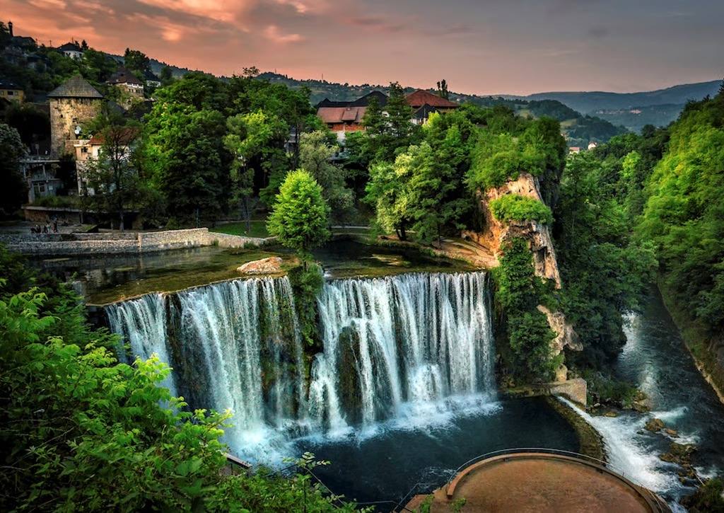 Idyllischer Pliva-Wasserfall in Jajce bei Sonnenuntergang, umgeben von grüner Natur, Felsen und traditioneller Architektur.