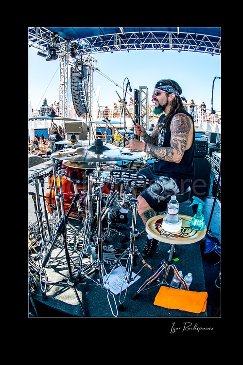 Vertical color image of Mike Portnoy playing drums on an outdoor stage in a wide fisheye concert scene with bright daylight and the crowd behind him