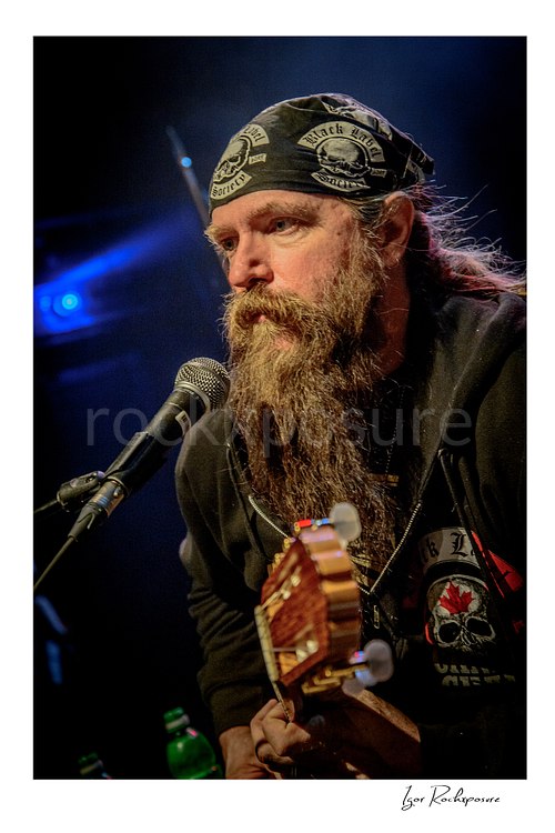 Close-up vertical shot of Zakk Wylde on stage during soundcheck, wearing a bandana and hoodie, holding an acoustic guitar next to a microphone.