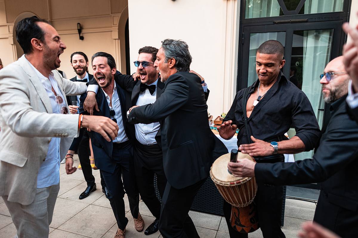 Rires et complicit&eacute; &eacute;clatent parmi un groupe d'hommes &eacute;l&eacute;gamment v&ecirc;tus, c&eacute;l&eacute;brant avec un percussionniste, dans une atmosph&egrave;re exub&eacute;rante et ensoleill&eacute;e, captur&eacute;e par Sebastien CLAVEL Photographe mariage Lyon