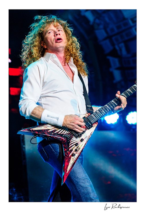 Vertical color image of Dave Mustaine of Megadeth playing guitar on stage with curly red hair and bright blue stage lights
