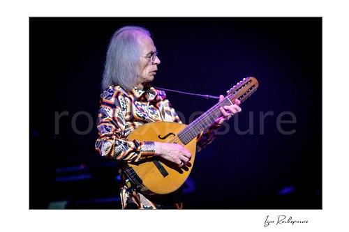 Horizontal color image of Steve Howe of Yes playing a small acoustic guitar onstage under purple concert light
