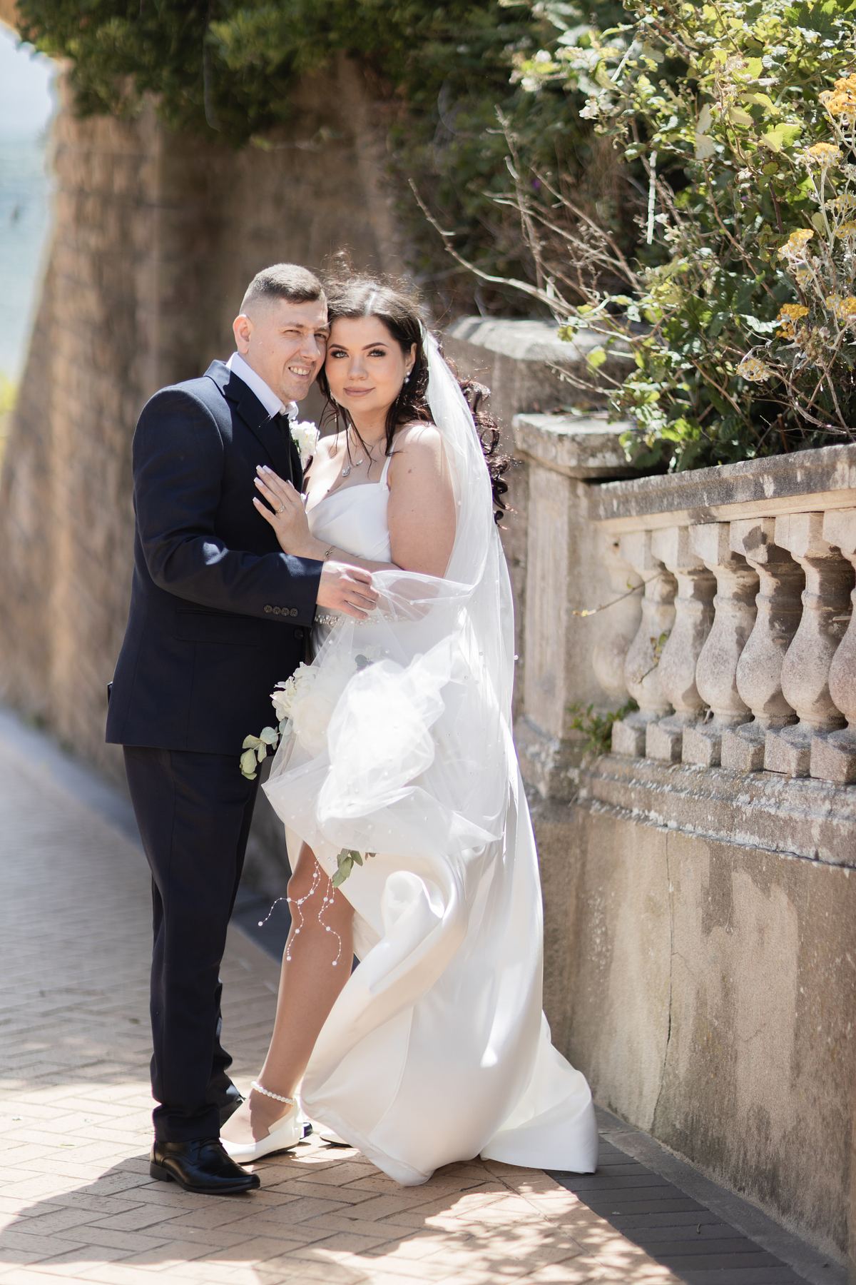 Bride and groom embracing beside a stone wall near the seafront at Barry Island, South Wales