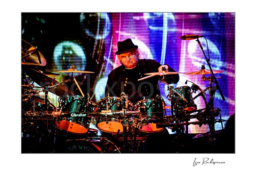Horizontal color image of Alan White of Yes performing at the drum kit before a vivid psychedelic stage screen