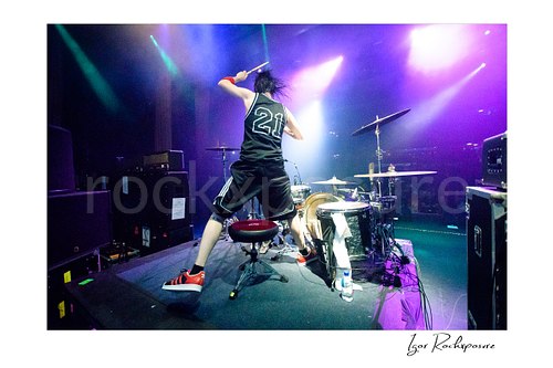 Horizontal color image of Zoltan Chaney performing on drums from behind on stage under purple and blue concert lighting