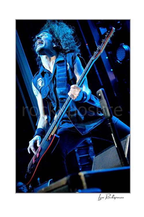 Vertical color image of Frank Bello of Anthrax performing live on stage, playing bass guitar under vivid blue concert lighting