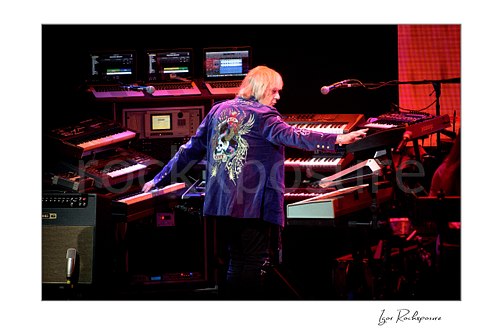 Horizontal color image of Geoff Downes of Yes performing at a large keyboard rig surrounded by multiple synthesizers during a live performance