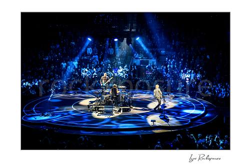 Horizontal color image of Muse performing live on a circular stage under vivid blue spotlights before a packed arena crowd