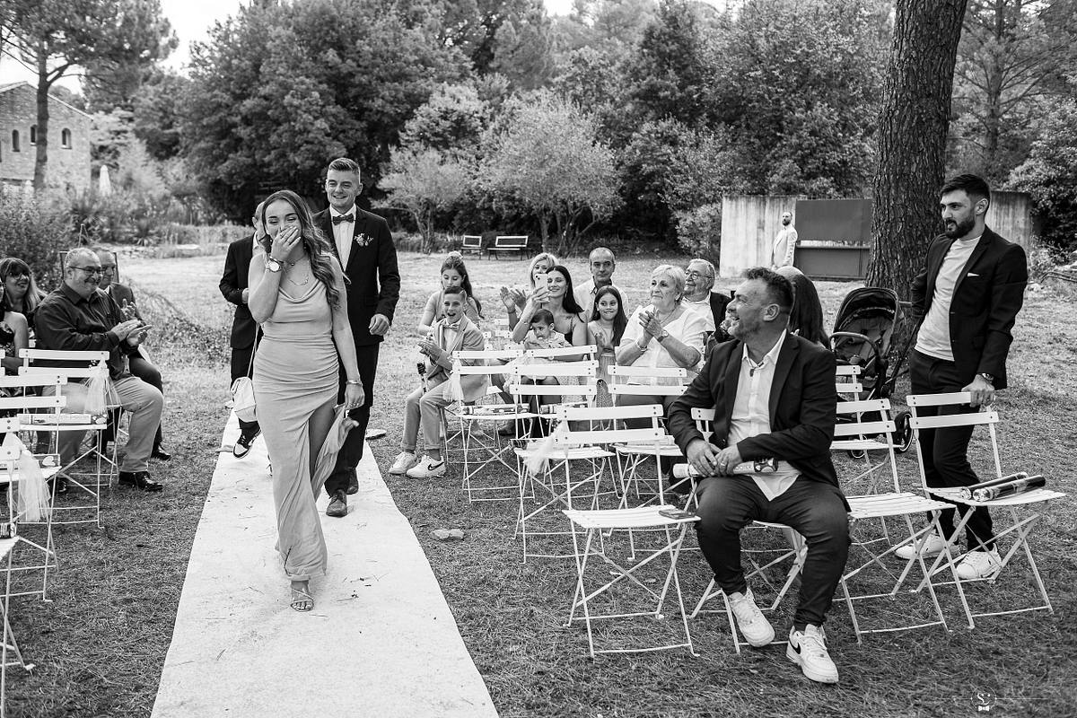 Un moment captivant de la c&eacute;r&eacute;monie la&iuml;que, o&ugrave; une invit&eacute;e &eacute;mue essuie une larme en traversant l'all&eacute;e, applaudie par les convives, et o&ugrave; se distinguent les sourires et la complicit&eacute; des proches pendant le mariage &agrave; N&icirc;mes