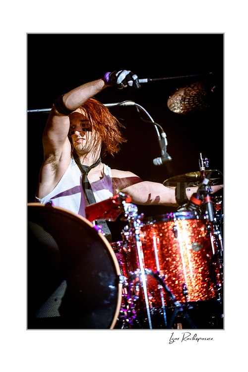 Vertical color image of Arejay Hale of Halestorm playing drums during a live performance, with one drumstick raised high above a sparkling red drum kit against a dark stage background