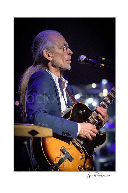 Vertical color image of Steve Howe of Yes playing guitar beside a microphone under cool purple concert light