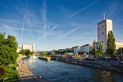 Weitwinkel-Aufnahme des Wiener Donaukanals im Sommer mit der bunt bemalten Uferpromenade und dem markanten Ringturm im Abendlicht unter blauem Himmel mit Kondensstreifen.