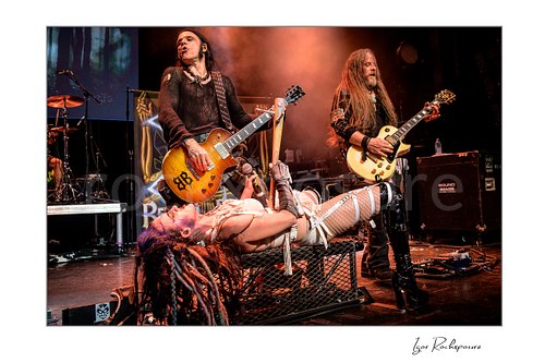 Horizontal color image of Chuck Garric, Calico Cooper, and Chris Latham of Beasto Blanco performing live on stage, with Cooper lying across a platform holding a microphone while Garric and Latham play guitar under warm stage light and smoke