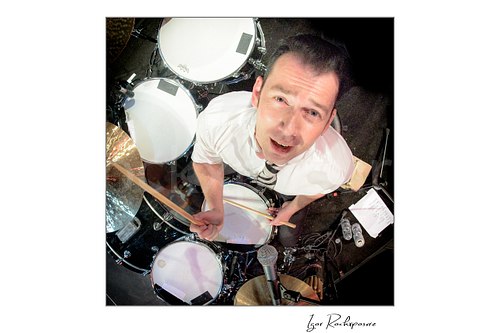 Square color image of Laust Sonne in an aerial overhead drummer shot during a live performance, looking up toward the camera with drumsticks in hand and the full kit visible