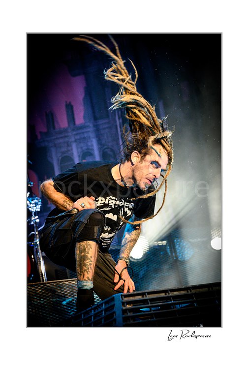 Vertical color image of Randy Blythe of Lamb of God crouched on stage with dreadlocks flying upward and dramatic backlight