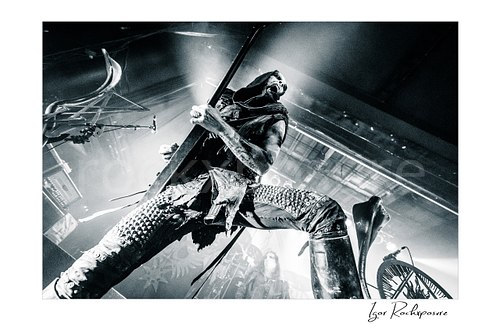 Horizontal black and white image of Nergal of Behemoth performing with a guitar in a dramatic low-angle stage shot under intense backlight