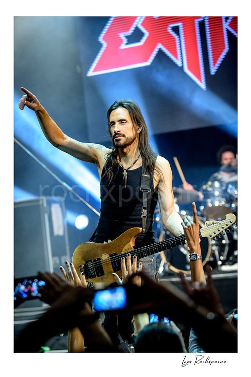 Vertical color image of Nuno Bettencourt performing live on stage with an electric guitar, pointing outward above the crowd