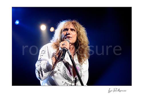 Horizontal color close-up image of David Coverdale of Whitesnake performing live on stage, singing into a microphone while holding a mic stand under blue concert lighting