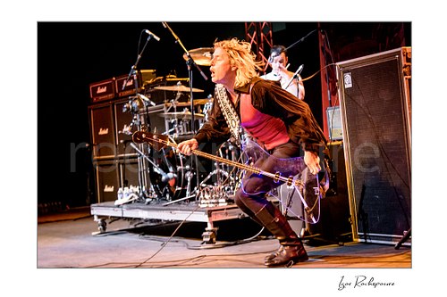 Horizontal color image of Stig Pedersen crouching low on stage during a live performance, with his bass guitar extended forward and Laust Sonne visible behind the drum kit